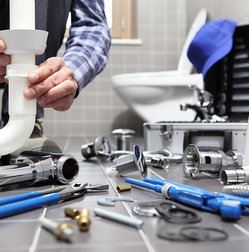 Plombier assemblant une tuyauterie de lavabo dans une salle de bain carrelée. Des outils, robinetterie et pièces sont au sol pour installation.