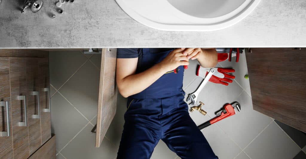 Plombier en salopette bleue sous un évier de salle de bain, réparant la plomberie avec des outils. Gants et clés à molette sur le carrelage.
