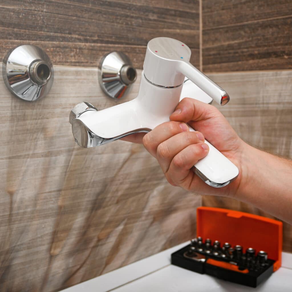 Main tenant un nouveau robinet mitigeur blanc et chromé devant des raccords muraux. Un kit d'outils est visible. Installation de robinetterie de salle de bain.