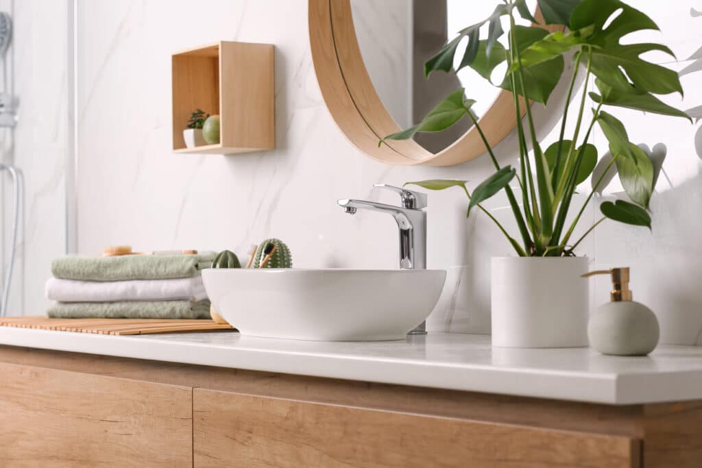 Salle de bain moderne: lavabo vasque, robinet chromé, miroir rond bois. Comptoir marbre, meuble bas bois. Serviettes, plantes.